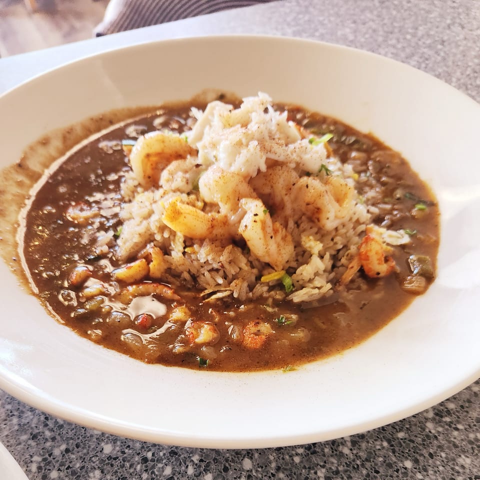 A white bowl filled with dark brown gumbo, rice, shrimp, and crab meat.