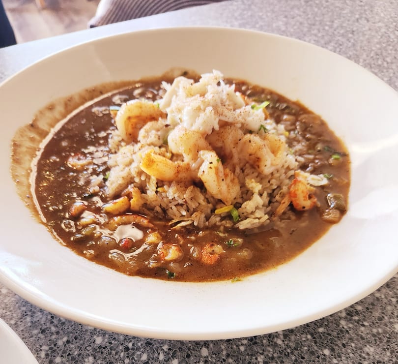 A white bowl filled with dark brown gumbo, rice, shrimp, and crab meat.